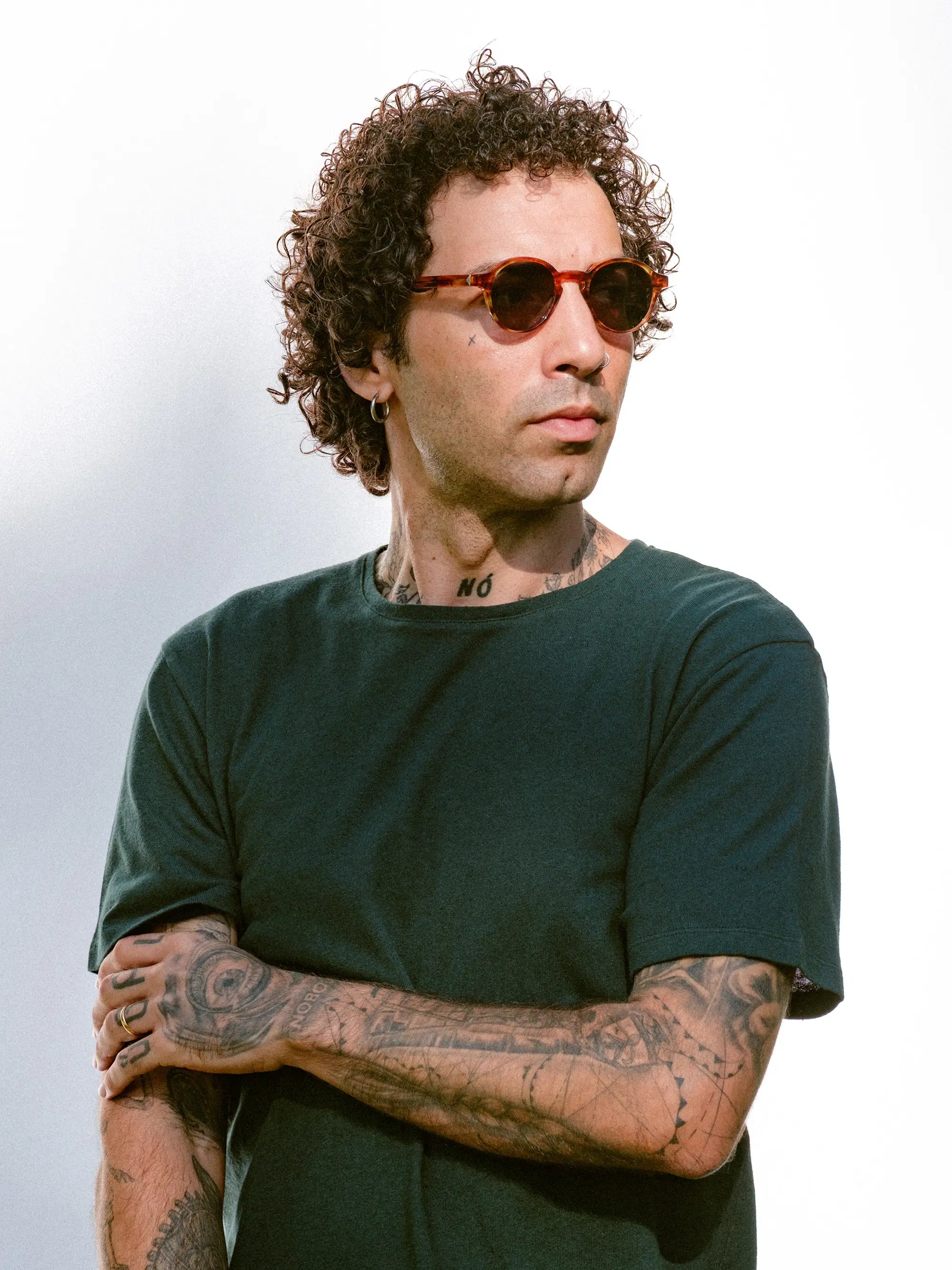 Man wearing sunglasses and a dark green t-shirt with tattoos on arms against a light background
